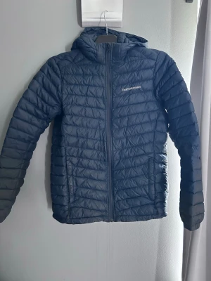 Svart dunjacka från Peak Performance - Snygg mörkblå dunjacka från Peak Performance med huva och dragkedja framtill. Jackan har quiltat mönster och två sidofickor med dragkedja. Perfekt för kalla vinterdagar och enkel att matcha med olika outfits. Storlek 170