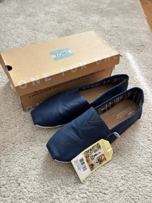 Mörkblå espandrillos från TOMS - Snygga mörkblå loafers från TOMS i textil med klassisk slip-on design. Skorna har en vit sula och ett färgglatt mönstrat innerfoder. Perfekta för dig som gillar enkel och stilren look. Levereras med originalkartong och tag kvar.