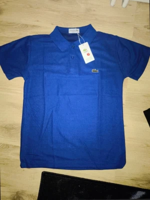 Lacoste Regular Fit Polo T-shirt- Marinblå (ny med taggar) - Helt ny Lacoste pikétröja i klassisk marinblå färg. Skick: Helt ny, aldrig använd. Passform: Regular Fit (bekväm, klassisk passform).  Material: Bomull.  Detaljer: Klassisk krage, två knappar framtill och den ikoniska broderade krokodilen på bröstet.  Färg: Marinblå. Mått: Axelbredd: ca 44 cm .           Längd: ca 62 cm . Perfekt för både vardag och finare tillfällen. Säljes då den inte kom till användning.