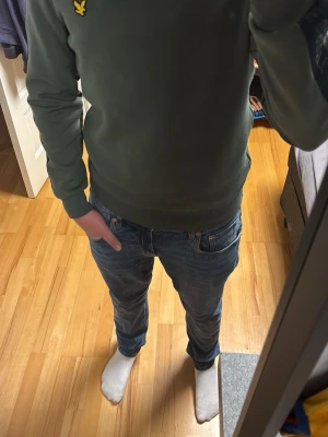 Mossgrön sweatshirt från Lyle & Scott - Mossgrön sweatshirt från Lyle & Scott med svart logotypband längs ärmarna och klassisk gul logga på bröstet. Tröjan har rund halsringning, muddar vid ärmslut och söm vid nederkant samt en dragkedja på sidan. Perfekt för en avslappnad stil.