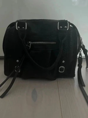 Svart handväska med dragkedjedetaljer - Cool svart handväska i boxig modell med silverfärgade metalldetaljer och dragkedja framtill, samt dekorativa remmar. Perfekt för dig som gillar en stilren men edgy look. Finns lite slitage på handtagen men är inget som syns. 