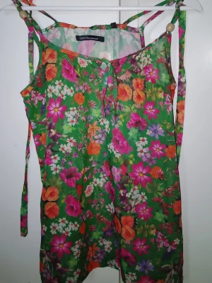 Blommig grön linnetopp från Emoi - Färgglad lite längre linnetopp från Emoi med grönt som bas och massor av blommor i rosa, orange, lila och vitt. Tunt och luftigt material, smala axelband med pärldetaljer och även ett snöre som följer med. Perfekt för varma dagar och sommarvibbar.