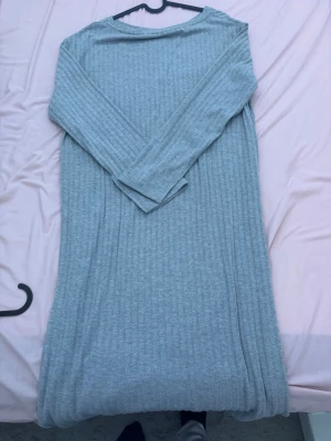 Ljusgrå ribbad långklänning - En stilren ljusgrå långklänning med ribbad struktur och långa ärmar. Klänningen har en rak passform och är perfekt för en enkel och avslappnad look. Tillverkad i mjukt material som känns skönt mot huden.