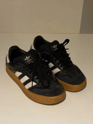 Adidas Samba OG svarta sneakers - Klassiska Adidas Samba OG sneakers i svart läder med vita detaljer och de ikoniska tre ränderna på sidorna. Skorna har svart snörning, vit hälkappa och en brun gummisula som ger en retrovibe. Perfekta för dig som gillar streetstyle och tidlös design.