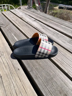 Burberry slides med klassiskt rutmönster - Snygga slides från Burberry med svart sula och bred rem i det ikoniska beigea rutmönstret med röda och svarta detaljer. Perfekta för sommaren och riktigt trendiga att bära till både shorts och jeans. Remmen är i syntetmaterial och sulan är mjuk och bekväm.