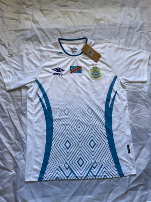 Vit DR Kongo fotbollströja Umbro - Snygg vit fotbollströja från Umbro med DR Kongos flagga och FECOFA-emblem på bröstet. Tröjan har blå grafiska mönster och detaljer, rund hals med blå kant och är tillverkad i lätt och ventilerande material. Perfekt för fotboll eller träning.