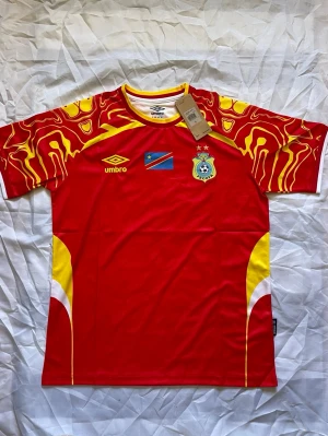 Röd Kongo fotbollströja från Umbro - Snygg röd fotbollströja från Umbro med gula och vita detaljer, samt mönstrade ärmar. Tröjan har Kongo-Kinshasas flagga och FECOFA-emblem på bröstet. Tillverkad i lätt och ventilerande material, perfekt för träning eller match.