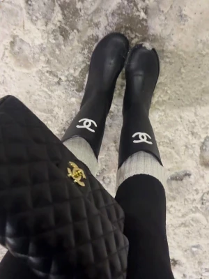 Chanel boots - Säljer ett par svarta stövlar från Chanel med klassisk vit logotyp framtill. Stövlarna är höga och har en stilren, minimalistisk design i gummi som passar perfekt för regniga dagar. De har en rund tå och grov sula för extra komfort och grepp.