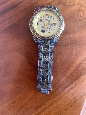 Vintageinspirerad skelettklocka i metall - Unik armbandsklocka med skeletturverk och detaljerat mönstrad länk i silverfärgad metall. Urtavlan är guldig med synliga kugghjul och gnistrande stenar som index. Klockan har en klassisk rund form och ett riktigt iögonfallande design.