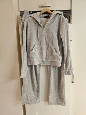 Grått mjukisset med luvtröja - Supermysigt grått mjukisset med zip-hoodie och matchande byxor. Hoodien har dragkedja, fickor och snören i huvan. Byxorna har resår i midjan och raka ben. Perfekt för chill dagar hemma eller när du vill vara bekväm. Materialet känns mjukt och skönt mot huden.