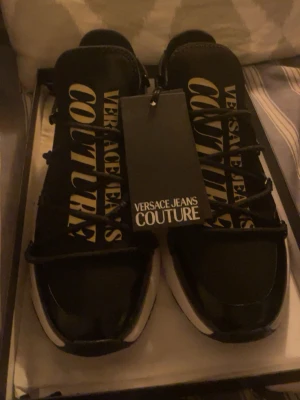 Svarta Versace Jeans Couture sneakers - Svarta sneakers från Versace Jeans Couture med stora vita logotexter på plösen och sidan. Skorna har en chunky vit sula och svarta snören. Snygg och modern design som sticker ut. Materialet är en mix av textil och syntet. 