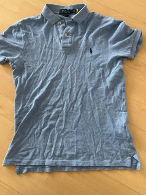 Blå pikétröja från Polo Ralph Lauren - Klassisk ljusblå pikétröja från Polo Ralph Lauren med krage, korta ärmar och knappar framtill. Slim fit-modell med broderad logga på bröstet. Perfekt för en stilren och avslappnad look.