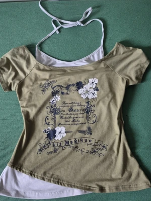 Grön topp med tryck och knytband - Trendig Grön topp med korta ärmar och vitt linne under, dekorerad med blommigt tryck och text framtill. Toppen har knytband i nacken och en lätt offshoulder-design som ger en cool look.