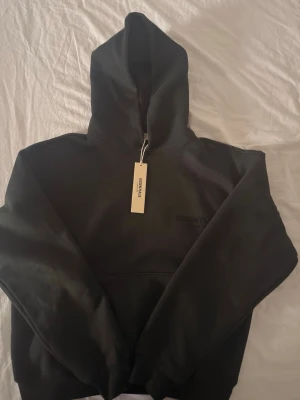 Svart Essentials hoodie med huva - Snygg svart hoodie från Essentials med broderad logga på bröstet och ryggen. Klassisk oversized passform, stor huva och känguruficka framtill. Tillverkad i mjukt och tjockt material som känns riktigt skönt. Perfekt för en avslappnad streetwear-look.