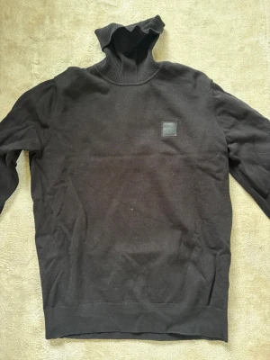 Svart polotröja från BOSS - Svart stickad polotröja från BOSS med diskret logotyp på bröstet. Tröjan har lång ärm, ribbstickad polokrage samt muddar vid ärmslut och nederkant. Perfekt för en stilren och klassisk look. Använts 1 gång sedan bara legat i garderoben, därav är den skrynklig. 