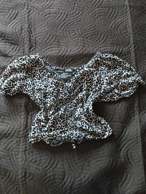 Leopardmönstrad croppad topp - Snygg croppad topp med leopardmönster i svart och beige. Toppen har puffiga korta ärmar och smockad midja som ger en trendig siluett. Perfekt för dig som vill sticka ut lite extra. Ganska kort i storleken men annars bra!