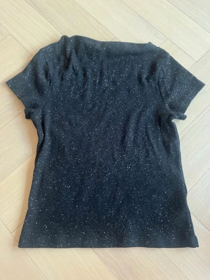 Svart glittrig T-shirt  - Svart t-shirt med korta ärmar och diskret ribbad struktur. Prickigt mönster i vitt ger en cool och avslappnad vibe. Perfekt till jeans eller kjol för en enkel men snygg look.
