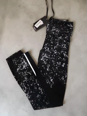 Svarta mönstrade leggings från Under Armour XS - Snygga svarta leggings från Under Armour med grått och vitt abstrakt mönster. De har hög midja, perforerade detaljer längs benen och en vit rand på ena benet. Tillverkade i stretchigt funktionsmaterial som passar perfekt till träning eller chill.