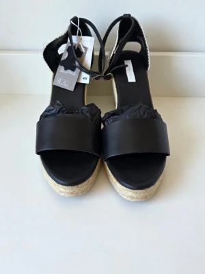Svarta kilklackade sandaler H&M - Snygga svarta sandaler från H&M med bred rem över foten och kilklack i flätad jute. Sandalerna har öppen tå och justerbar rem runt ankeln. Perfekta för sommaren och ger en trendig look till alla outfits.