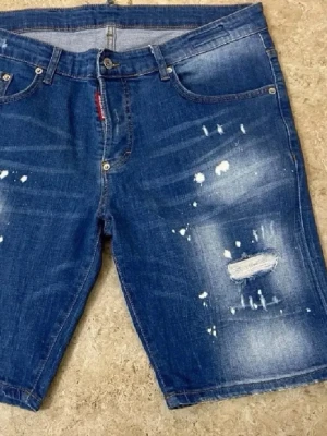 Blå denimshorts med slitningar - Snygga blå denimshorts med slitna detaljer och ljusa partier framtill. Klassisk femficksmodell med knapp och dragkedja. Perfekta för en avslappnad och trendig look.