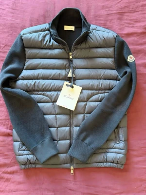 Moncler cardigan - Snygg grå dunväst från Moncler med quiltad front och stickade ärmar. Västen har hög krage, dragkedja framtill och två sidofickor med dragkedja. Klassisk Moncler-logga på ärmen och elastisk nederkant för skön passform. Perfekt för lager på lager och streetstyle.