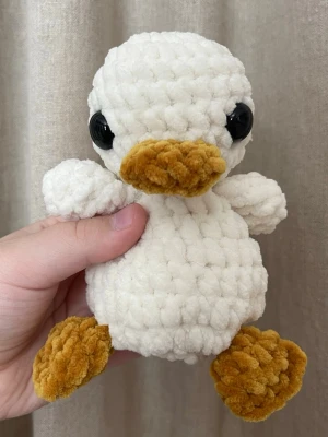 Virkad anka i vitt – varm ton – handgjord plushie - Handvirkad anka i vitt med varmare beige/guldiga detaljer. Mjuk och stilren, passar både barnrum och som söt inredning. Varje exemplar är unikt och gjort för hand. Storlek: ca 15 cm.