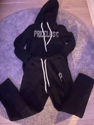 Svart  Priceless Tracksuit - Svart loungewear-set med hoodie och byxor från Priceless. Hoodien har vit dragsko och tryck i vitt framtill. Byxorna har matchande vit dragsko och ett litet 'P' tryckt på ena benet. Perfekt för chill dagar hemma.