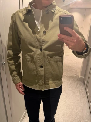 Beige overshirt från J.Lindeberg - Stilren grön/beige overshirt från J.Lindeberg med klassisk krage och stora frontfickor. Jackan har raka linjer, knäppning framtill med fyra knappar och är tillverkad i ett kraftigare bomullstyg. Perfekt för lager-på-lager och enkel att matcha.