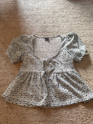 Blommig blus med puffärm och snörning - Supersöt blommig blus i vitt och blått med puffärmar och snörning framtill. Blusen har en lätt v-ringad urringning och volang nedtill. Perfekt för sommarens alla tillfällen. 