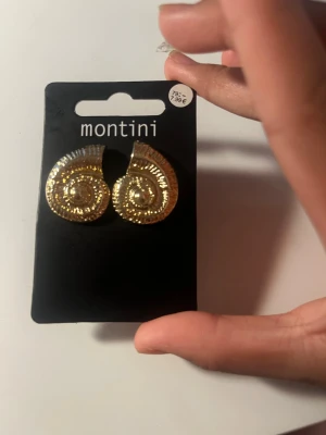 Guldiga snäckformade örhängen Montini - Säljer ett par coola örhängen från Montini i guldig metall med snäckformad design och detaljerad yta. Perfekta för att ge din look lite extra edge och glans. Säljs på originalkortet.