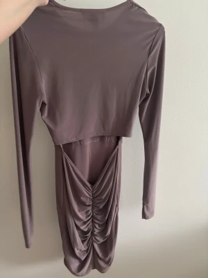 Ljuslila/brun långärmad klänning med rynk - Trendig ljuslila/brun klänning från Rebecca Stella med lång ärm och rund halsringning. Klänningen har en snygg rynkad detalj baktill, öppen i rygg. Tillverkad i mjukt, stretchigt material som sitter bekvämt på kroppen. Passar till både xs och S. Klänningen slutar vid knäna.