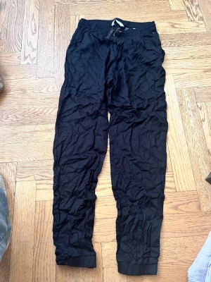Svarta mjukisbyxor från H&M - Svarta mjukisbyxor från H&M med resår och dragsko i midjan. Byxorna har muddar vid bensluten och är tillverkade i ett mjukt och lätt material. Perfekta för en avslappnad stil och riktigt sköna att ha på sig.