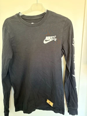 Svart långärmad tröja från Nike - Svart långärmad tröja från Nike med tryckt logga på bröstet och coola vita detaljer på ärmarna. Rund halsringning och patch med 'Nike Sportswear' nertill. Perfekt för en avslappnad streetstyle-look.