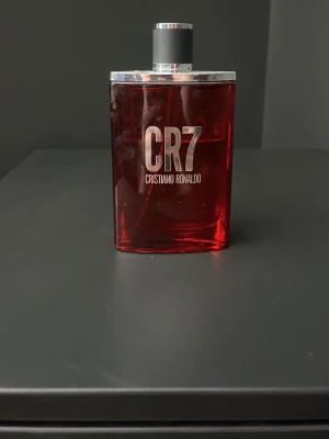 CR7 parfym av Cristiano Ronaldo - Snygg parfymflaska i rött glas med silverdetaljer från Cristiano Ronaldo, CR7. Flaskan har en stilren, rektangulär form och ett lock med en siluett av Ronaldo. Perfekt för dig som gillar sportiga och fräscha dofter. Det är ungefär 70 ml kvar i flaskan. Bara att skriva vid frågor!