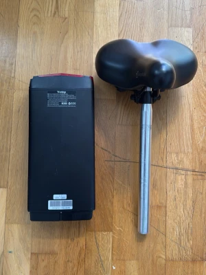 Cargobike Elsparkcykelbatteri & sadel - Säljer ett svart batteri till elsparkcykel av märket Wangja samt en svart bred sadel med silverfärgat sadelrör. Batteriet har handtag och knappar på ovansidan. Perfekt för dig som behöver reservdelar till din elsparkcykel.