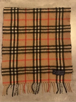 Burberry halsduk - Klassisk beige halsduk från Burberrys i ikoniskt rutmönster med svarta, vita och röda ränder. Halsduken är tillverkad i en lyxig mix av 50% kashmir och 50% ull och har fransar i ändarna. Perfekt accessoar för att lyfta din outfit.