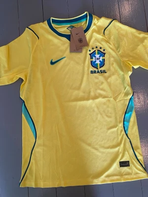 Brasilien Nike fotbollströja gul - Gul Brasilien fotbollströja från Nike med blå detaljer och CBF-emblem på bröstet. Tröjan har korta ärmar, rund halsringning med blå kant och är tillverkad i lätt, ventilerande material. Perfekt för fotbollsträning eller att bära när du hejar på landslaget.