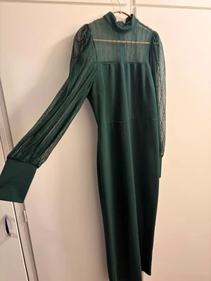 Grön långklänning med meshärmar - Stilren grön långklänning med höghalsad krage och transparenta meshärmar. Klänningen har långa ärmar med breda muddar och en elegant, rak siluett. Perfekt för dig som vill sticka ut med en unik detalj på ärmarna.