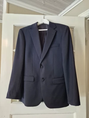 Mörkblå kostym med kavaj och byxor - Snygg mörkblå kostym med klassisk kavaj stl 48, väst stl S, slips och matchande kostymbyxor stl 30-30 Kavajen har två knappar, fickor med lock och en bröstficka. Byxorna har pressveck och bälteshällor. Perfekt för dig som vill ha en stilren och tidlös look. Endast använd till studenten 2025 av kille ca 175 cm smal kroppsbyggnad.