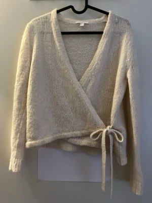 Beige omlottstickad tröja - Mysig beige stickad omlott-tröja med knytband i midjan och djup V-ringning. Långärmad och i mjukt, fluffigt material som ger en avslappnad och trendig look. Perfekt att slänga på sig över ett linne eller t-shirt.
