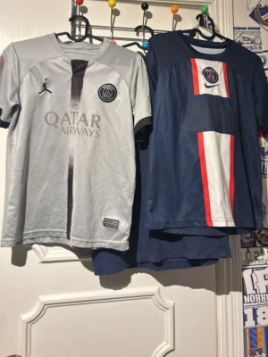 PSG Neymar Jr matchtröjor - Två snygga PSG matchtröjor med Neymar Jr och nummer 10 på ryggen. En är grå med svarta detaljer och Qatar Airways-logga, den andra är marinblå med röd och vit rand samt PSG-emblem. Perfekta för fotbollsfans som vill sticka ut på planen eller läktaren.