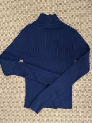 Mörkblå ribbstickad polotröja - Snygg mörkblå ribbstickad polotröja med lång ärm och hög krage. Tröjan har en figurnära passform och är tillverkad i ett mjukt, stretchigt material som passar perfekt till lager på lager. Enkel och stilren design utan synliga loggor.
