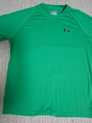 Grön t-shirt från Under Armour - Snygg grön t-shirt från Under Armour med klassisk rund hals och diskret logga på bröstet. Tillverkad i ett lätt och funktionellt syntetmaterial som andas, perfekt för träning eller chill. Modellen är loose fit för en avslappnad vibe.