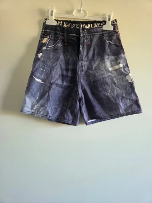 Blå denimshorts 122/128 - Snygga blå denimshorts från Street life med coola slitningar och röda detaljer på bakfickorna. Klassisk femficksmodell med knapp och dragkedja. Perfekta för en avslappnad stil under varma dagar.