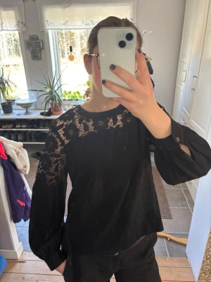 Svart blus med spetsdetaljer - Snygg svart blus med transparenta spetsdetaljer över axlar och armar. Blusen har rund halsringning och vida långärmade ärmar med knapp vid ärmslut. Perfekt för dig som vill ha en elegant och trendig look.