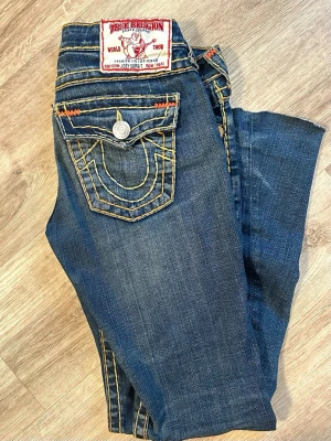 True Religion bootcut low waist jeans blå - Säljer ett par blå bootcut jeans från True Religion med fat stitch tjocka orangea sömmar och  detaljer på bakfickorna. Jeansen är lågmidjade och bootcut. Har lite slitage vid fötterna och en liten fläck vid benet. 🥰💗 kan skicka fler bilder och priset kan diskuteras 