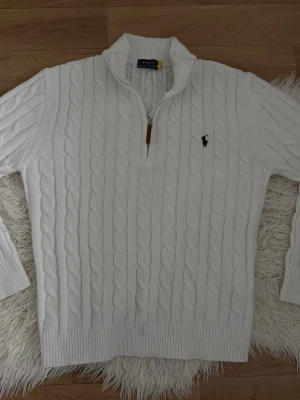 Vit kabelstickad tröja Polo Ralph Lauren - Vit kabelstickad tröja från Polo Ralph Lauren med halv dragkedja och hög krage. Tröjan har det klassiska broderade logotypen på bröstet och ribbade muddar. Perfekt för lager-på-lager och snygg till jeans.