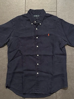 Mörkblå kortärmad  linneskjorta Ralph Lauren - Snygg mörkblå skjorta från Ralph Lauren med korta ärmar och klassisk button-down krage. Skjortan har vita knappar framtill och den ikoniska röda broderade loggan på bröstet.