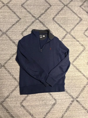 Blå halvzip från Polo Ralph Lauren storlek S - Mörkblå långärmad tröja från Polo Ralph Lauren med halvzip och grå insida på kragen. Klassisk röd logga broderad på bröstet. Perfekt för en stilren och avslappnad look. Storlek S