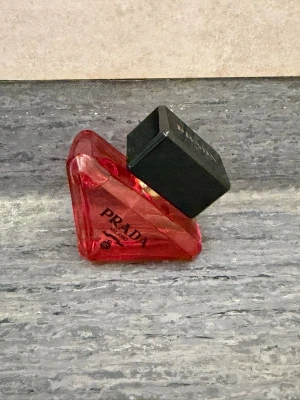 Prada Paradoxe Eau de Parfum - Unik parfymflaska från Prada i en cool, trekantig form. Flaskan är gjord av rött glas med svart lock och har märkets logga tryckt på framsidan. Perfekt för dig som gillar exklusiva och trendiga dofter.